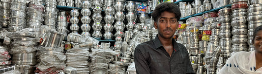 Man in a shop in India with various stainless steel products from India. Including pots, stainless steel plates, stainless steel cups