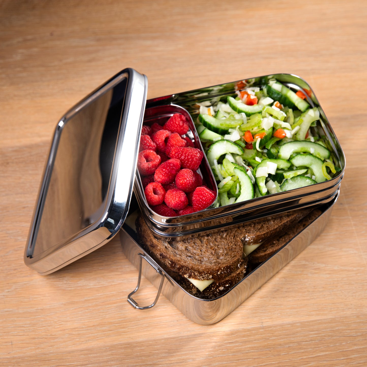 Stainless steel lunch box with compartments filled with lettuce, raspberry and bread. on a wooden surface