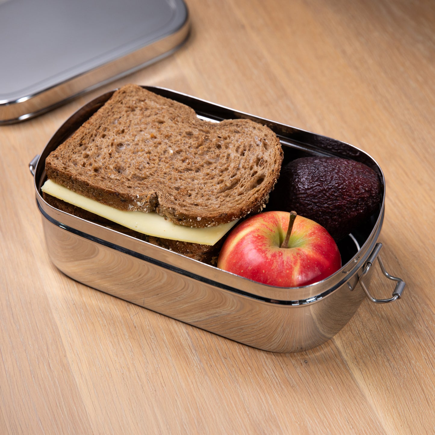 Stainless steel lunch box filled with bread and apple on a wooden surface with a lid on top