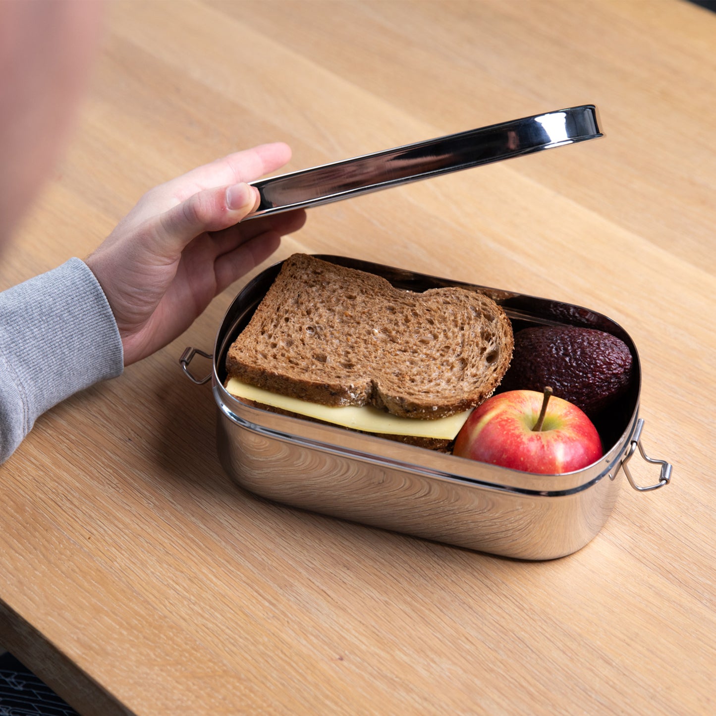 Stainless steel lunch box that is opened by one hand. The lunch box contains a sandwich and an apple