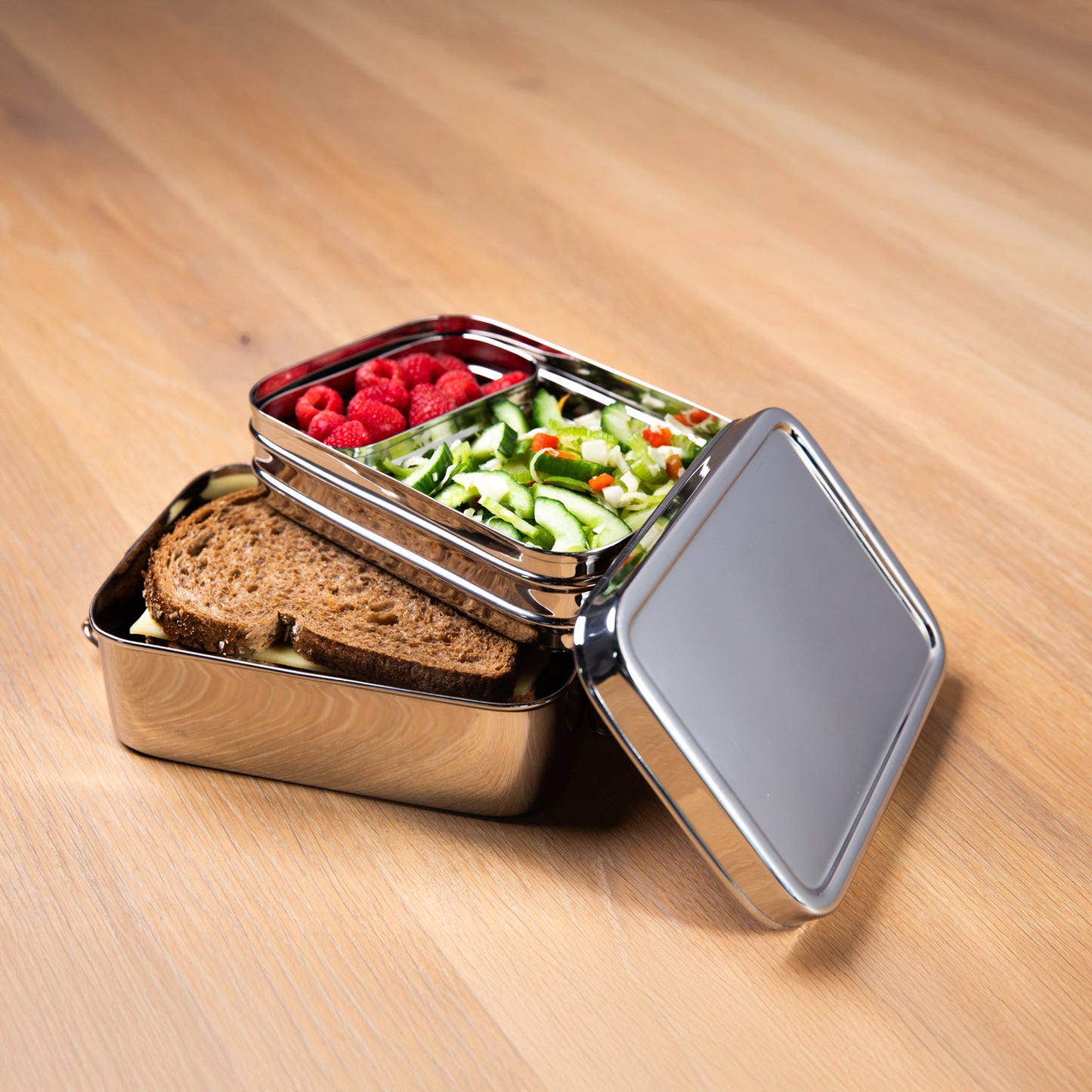 Stainless steel lunch box with compartments for adults filled with lettuce, fruit and bread. on a wooden surface