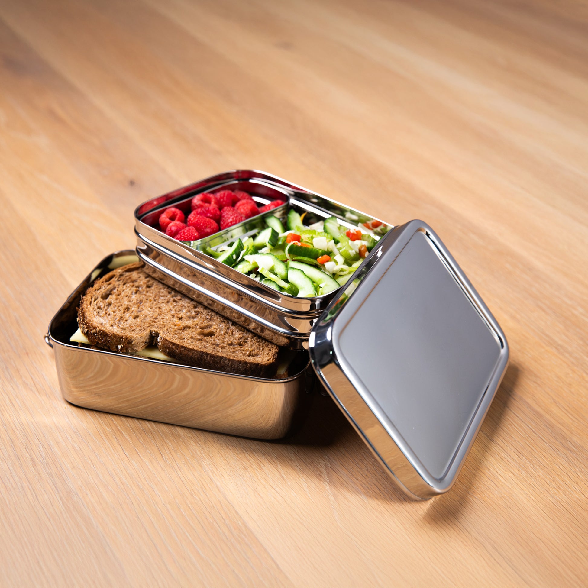 Stainless steel lunch box with compartments for adults filled with lettuce, fruit and bread. on a wooden surface