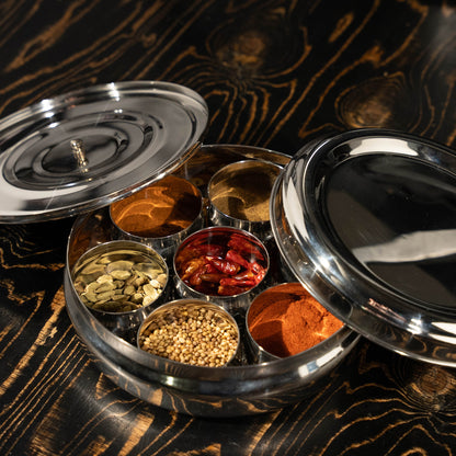 Stainless steel masala dabba filled with spices on a black table