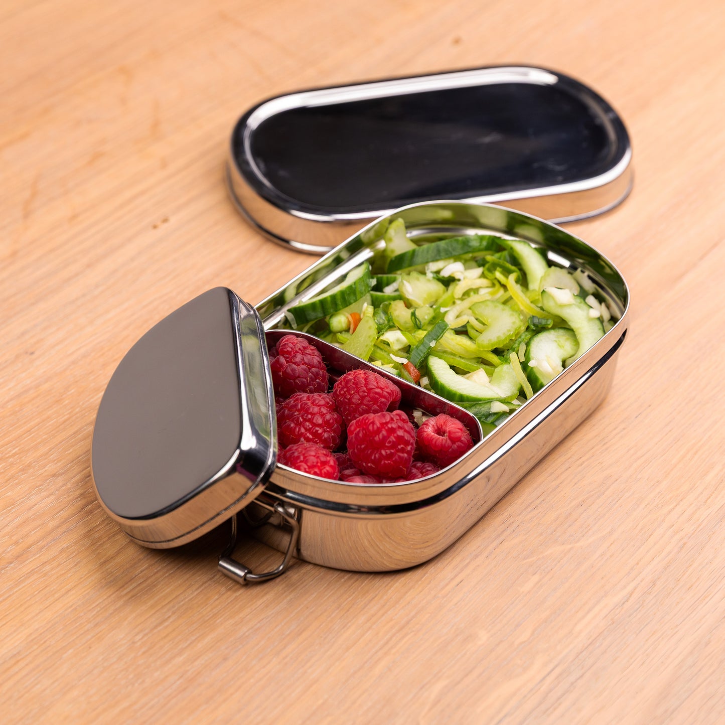 Stainless steel lunch box with compartments filled with raspberry and salad on a school table